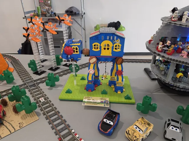 A LEGO AT-AT built in Fabuland style: blue body, yellow round windows, standing on a green baseplate beside Duplo vehicles and a grey Death Star