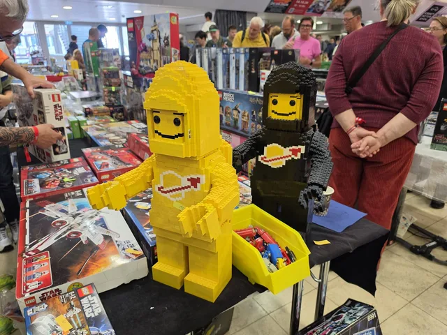 Two brick-built minifigures about knee height on a trader's table — a yellow one with the Classic Space logo and a black one — surrounded by boxed Star Wars sets